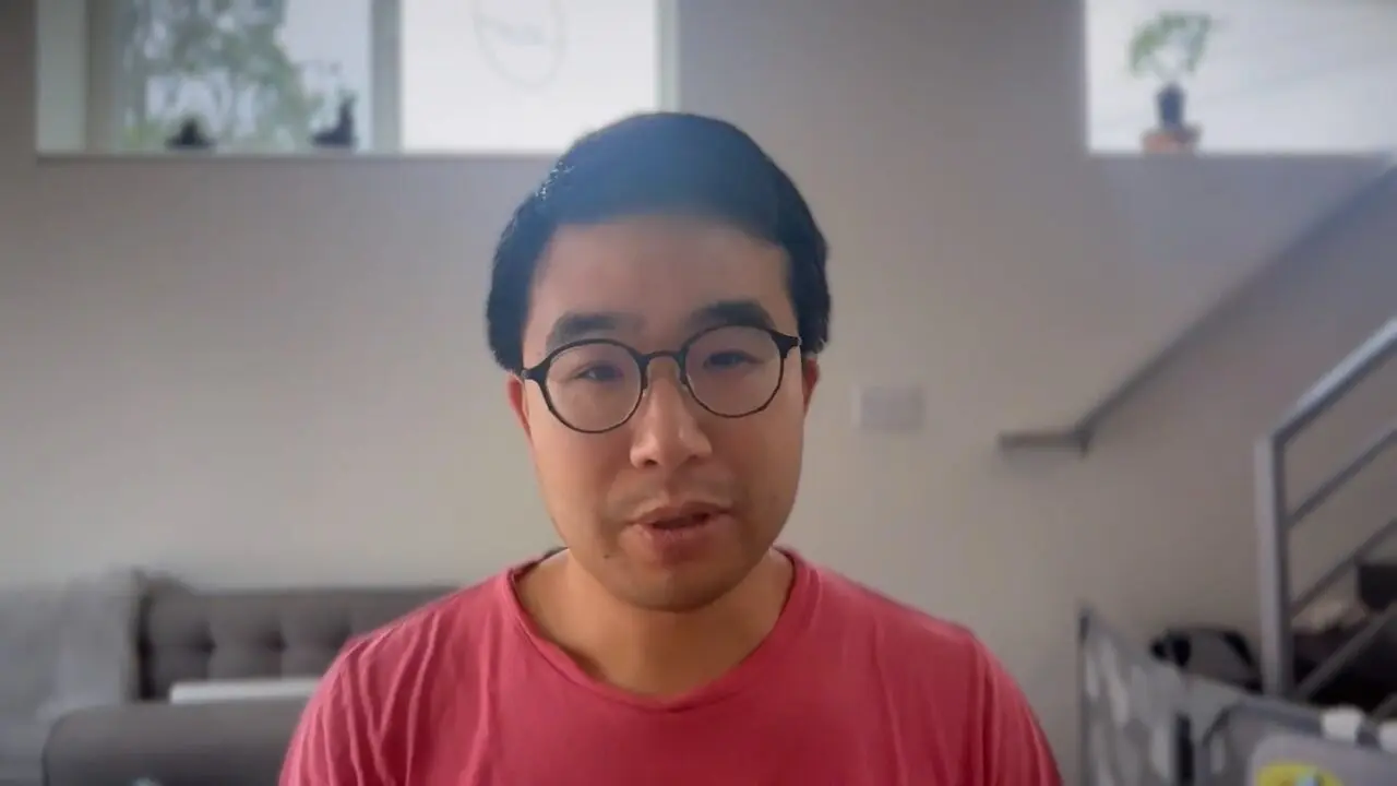 Person in glasses and pink shirt speaking in a cozy indoor setting.