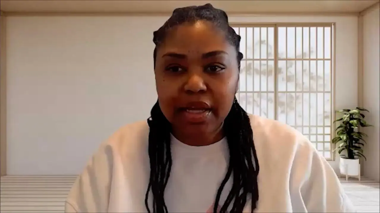 Person speaking on a video call, wearing a casual white top, with a minimalist room background.