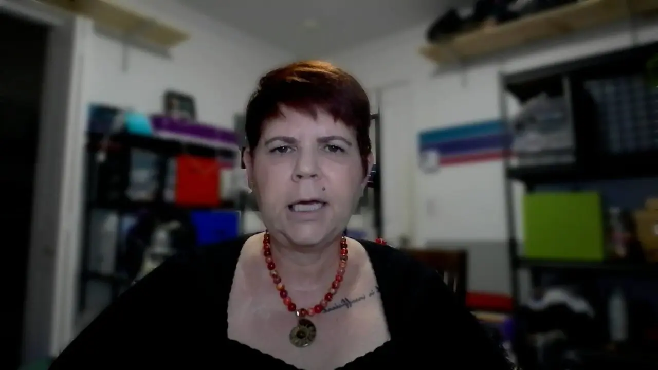 Person with red necklace speaks in a cluttered room, expressing emotion with hand gestures.