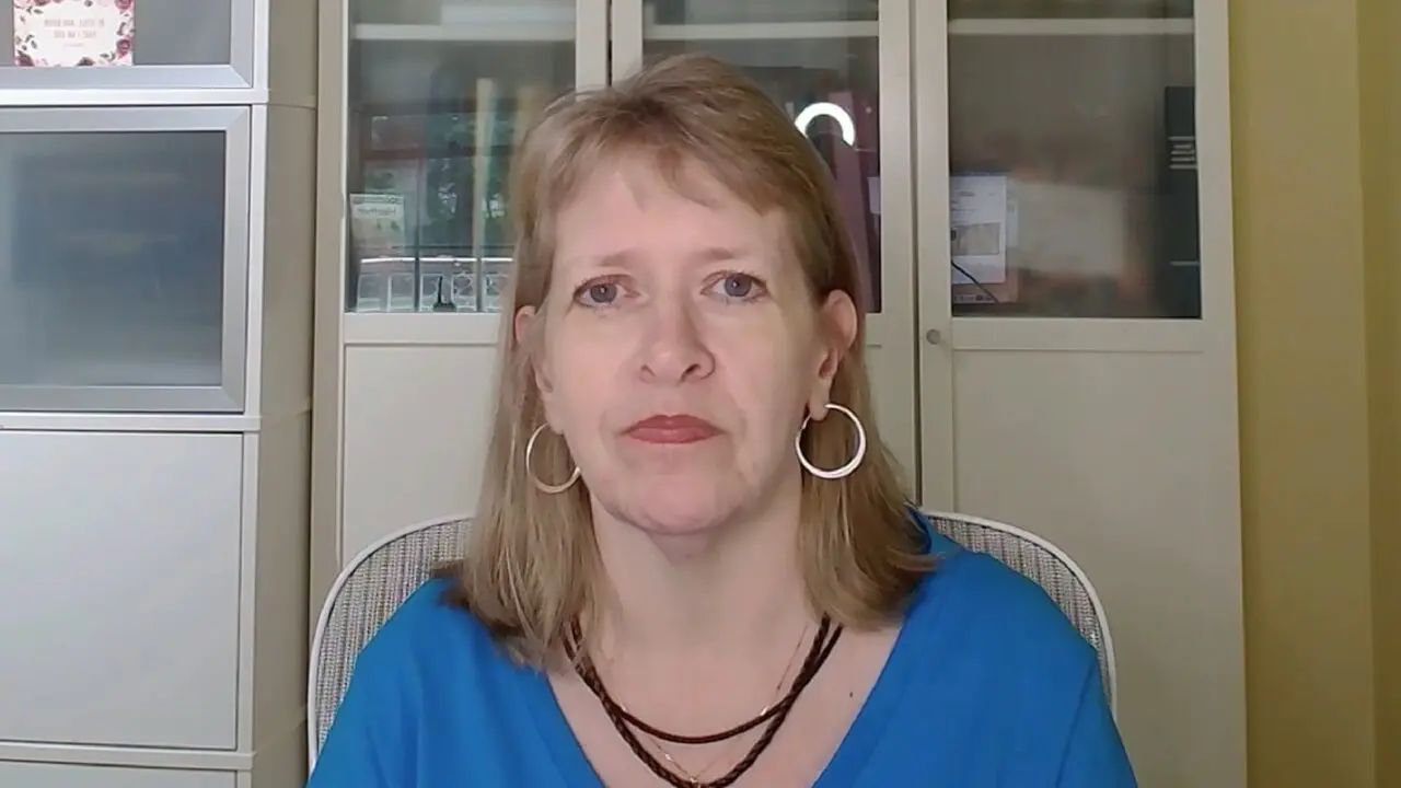 Neutral expression individual in blue shirt, hoop earrings, seated indoors with cabinet background.