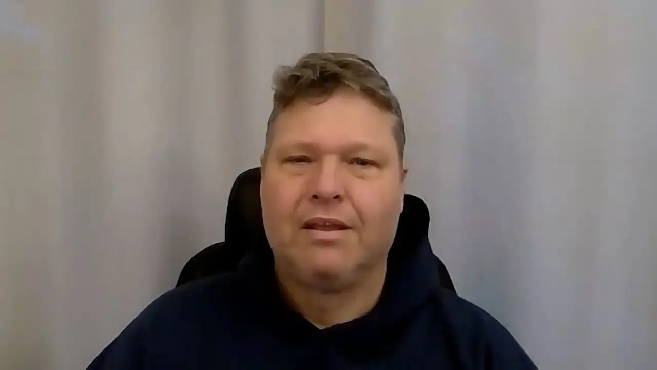 Person seated, wearing a dark hoodie against a plain curtain backdrop.