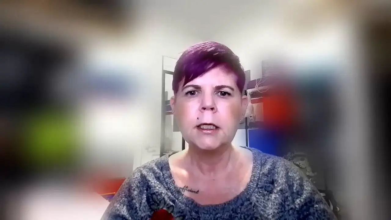 Person with short purple hair, wearing a sweater, in a colorful blurred background office.