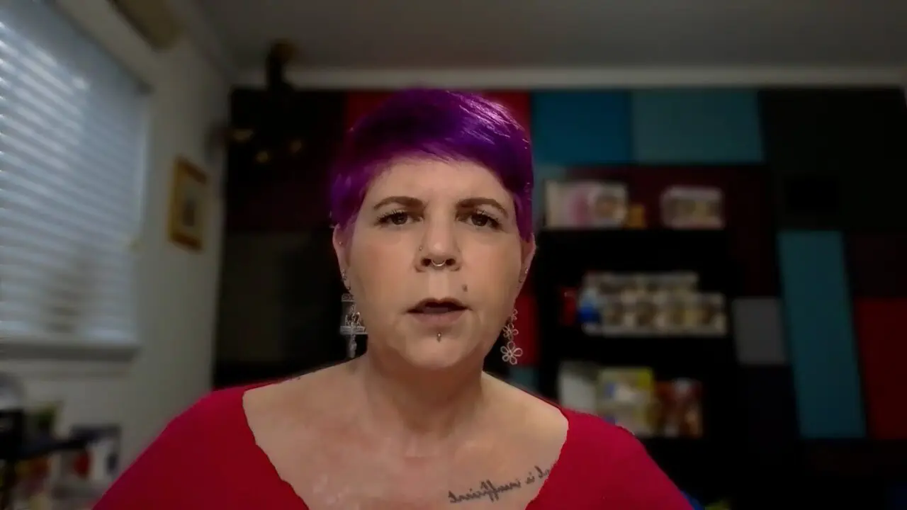 Person with vibrant purple hair speaks indoors, wearing earrings and a pink top.
