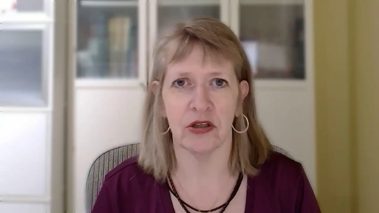 Person wearing hoops and layered necklace, sitting indoors against a blurred office background.