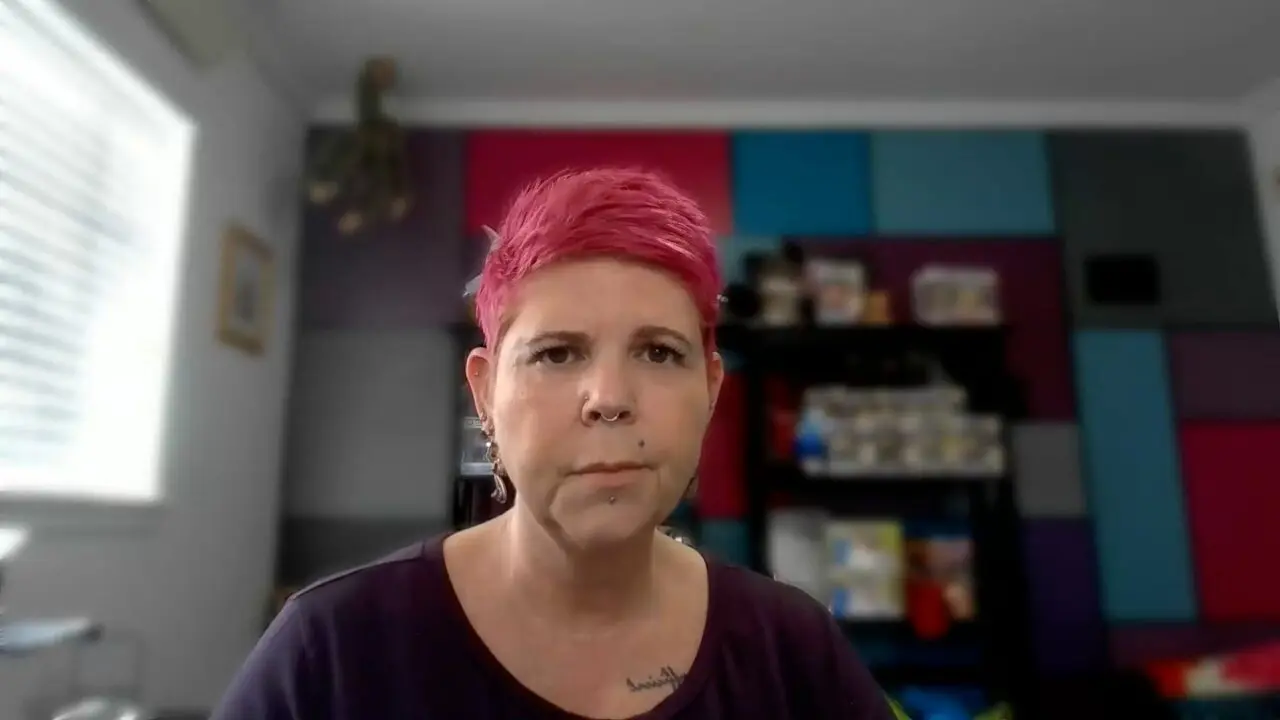 Person with pink hair in a colorful room, facing camera with neutral expression.