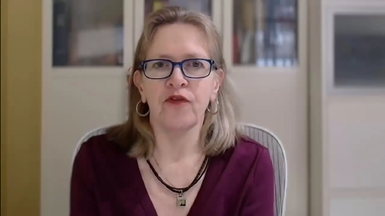 Person in glasses and earrings speaking, wearing a necklace and maroon top indoors.